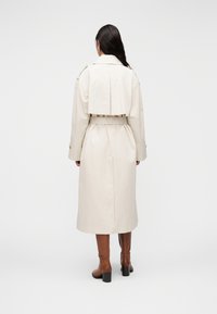 Μπεζ καμπαρντίνα με ζώνη στη μέση, διπλή στρώση στην πλάτη και μακριά μανίκια. Φοριέται πάνω από καφέ μπότες μέχρι το γόνατο με τετράγωνες φτέρνες.