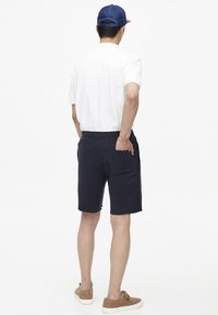 Pantaloni corti blu in cotone con tasche laterali, abbinati a una maglietta bianca. Il modello indossa un cappellino blu e scarpe marroni.