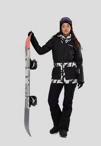 Zwarte sneeuwjack met witte patroonzakken, gecombineerd met zwarte sneeuwbroek. Model houdt een snowboard vast met gedurfde graphics.