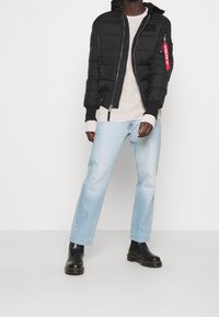 Person iført en sort vinterjakke, beige sweater, lyseblå jeans og sorte læderstøvler, der står foran en hvid baggrund.