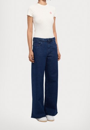 Osoba nosząca białą, krótko rękawową, teksturowaną bluzkę oraz ciemnoniebieskie, szerokie spodnie z wysokim stanem i białe tenisówki.