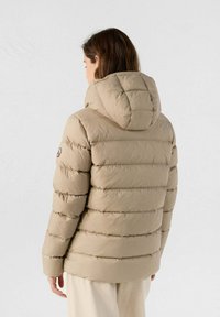 Frau in einer beigen, gesteppten Daunenjacke mit Kapuze, die sich von einer hellen, einfarbigen Hintergrund abwendet.