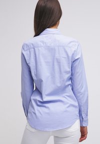 Camicia a maniche lunghe di color azzurro chiaro con colletto, texture liscia, polsini con bottone e orlo curvo, caratterizzata da un design elegante e su misura.