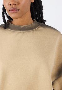 Beige sweatshirt met een hoge hals, zachte textuur en een gradueel donkerdere tint bij de schouders. Het heeft subtiele stiksels.