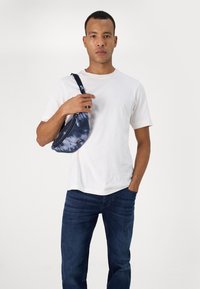 Modrý batoh přes rameno s tie-dye vzorem, vyrobený z textilu, ve tvaru půlměsíce, se zipovým uzávěrem. Modelka má na sobě bílý tričko a tmavé džíny, stojí na jednobarevném pozadí.