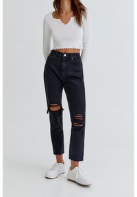 Black high-waisted jeans with distressed knee rips, paired with a cropped white long-sleeve top featuring a scalloped hem and v-neck.