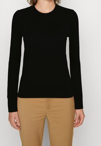 Femme portant un pull noir à manches longues ajusté et un pantalon beige, debout les bras détendus le long du corps sur un fond uni.