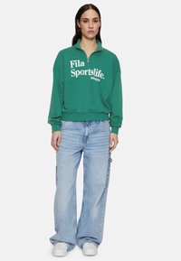 Felpa verde in poliestere con zip a metà, con testo "Fila Sportslife", abbinata a jeans blu chiaro a gamba larga, dotati di tasche laterali e sneaker bianche.