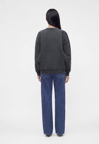 Personne aux longs cheveux bruns, de dos, portant un sweat-shirt noir délavé et un jean bleu ample avec des talons.