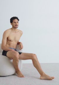 Fekete boxeralsó, szürke derékrész, egy texturált, világos színű kerek széken ül, sima fehér háttérrel. Nincsenek más figyelemre méltó részletek.