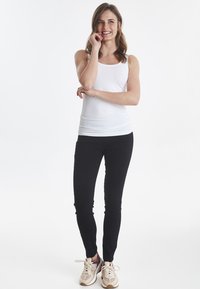 Weißes ärmelloses T-Shirt kombiniert mit schwarzen engen Hosen und hellen Sneakern. Das Model steht entspannt vor einem weißen Hintergrund.