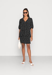 Vestido preto com bolinhas brancas, mangas curtas bufantes, decote em V, design em camadas e uma saia combinando. Acompanhado de sandálias brancas de dedo e uma bolsa texturizada.
