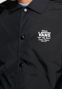 Giacca nera impermeabile con chiusura a bottoni a pressione. Presenta il logo Vans in bianco, un colletto classico e una texture liscia.