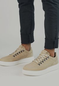 Sneakers in suede beige con suole in gomma bianca, dotati di occhielli in metallo e lacci rotondi. Pantaloni neri a sigaretta con risvolti.
