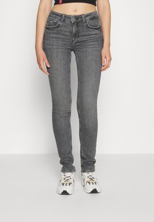 Czarne, wyblakłe jeansy o dopasowanym kroju, noszone z białymi i beżowymi sneakersami, widoczne od talii do stóp na jednolitym jasnym tle.