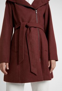 Bordowy wełniany płaszcz z paskiem w talii, kapturem i dwoma przednimi kieszeniami. Posiada zamek błyskawiczny oraz miękkie, teksturowane wykończenie.