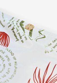 Biele tkanivo so zeleným kurzívnym textom, červenými kvetinovými vzormi a malou dekoratívnou zlatou ozdobou v strede.