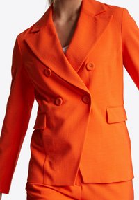 Vrouw in een feloranje dubbelzijdige blazer met bijpassende broek en een witte top eronder, tegen een witte achtergrond.