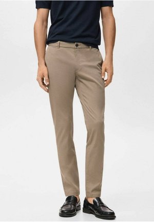 Mann, der beige Slim-Fit-Hosen, schwarze Loafers und ein dunkelblaues Hemd trägt, vor einem einfarbigen hellen Hintergrund steht.
