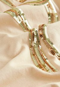 Beige tyg med vågiga rader av guld- och silverpaljetter som har en slät textur och ett lätt glansigt utseende, vilket skapar ett iögonfallande designelement.