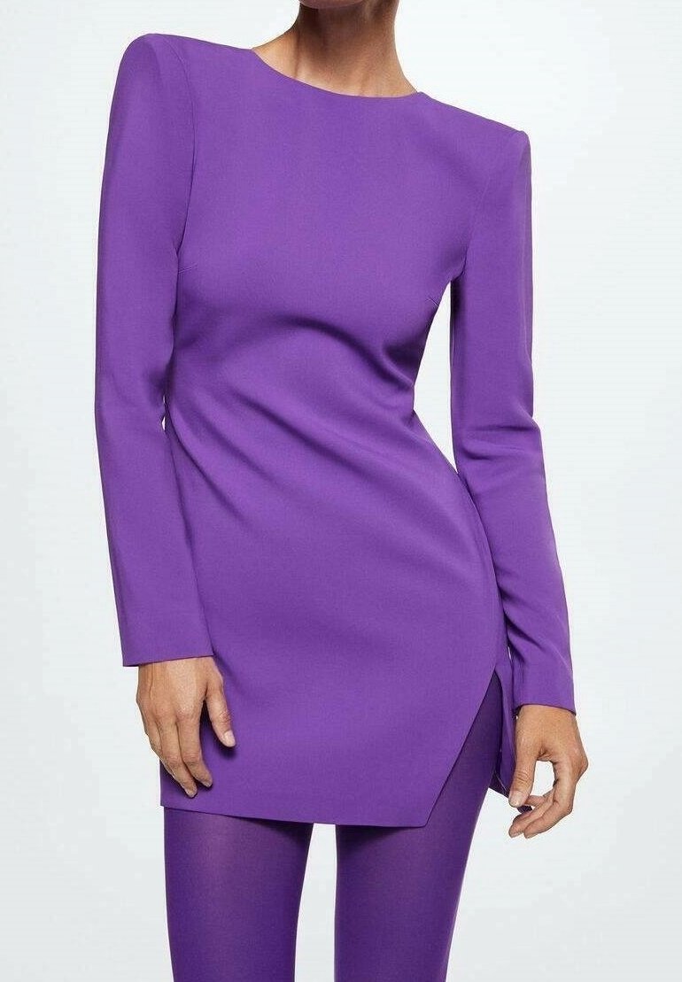 Femme portant un tunique asymétrique longue à manches, de couleur violette, associée à un legging violet assorti sur un fond clair et uni.