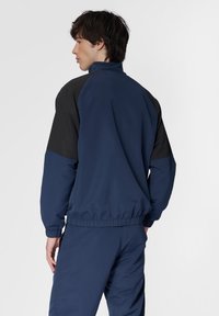 Mörkblå jacka med svarta axelaccenter, hög krage, elastiska ärmslut och midja. Slätt tyg med en avslappnad passform.