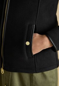 Ruka v vrecku čiernej fleecovej bundy so zlatým zipsom a patentkou, nosenej nad olivovými nohavicami.