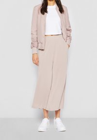 Donna che indossa pantaloni larghi beige, una giacca bomber abbinata, un top corto bianco e sneakers bianche, in piedi davanti a uno sfondo luminoso e semplice.