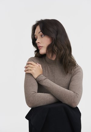 Haut à manches longues marron avec col roulé, au toucher lisse, avec une coupe ajustée et des détails de couture discrets. Le modèle est assis, les bras croisés, portant une bague en or.
