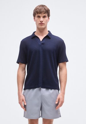 Sötét tengerészkék rövid ujjú galléros póló texturált mintával, gallérral és V-nyakkivágással, világosszürke rövidnadrággal viselve.