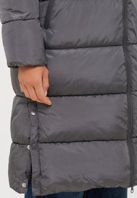 Giacca a vento grigia con una texture liscia, dettagli in bottoni sul orlo e chiusura con zip. Presenta sezioni trapuntate e maniche aderenti.