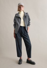 Grijze zip-up hoodie met een zachte textuur over een lichtgele coltrui, gecombineerd met donkerblauwe wijde broekspijpen en zwarte plateau loafers.