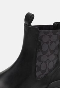 Schwarze Knöchelstiefel aus Leder mit einem strukturierten Stoffeinsatz, der ein sich wiederholendes Logomuster aufweist. Mit einer Zuglasche für einfaches Anziehen.