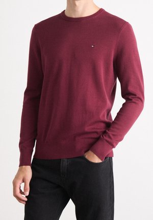 Uomo che indossa un maglione aderente bordeaux a girocollo con un piccolo logo sul petto, abbinato a jeans neri, con una mano in tasca.