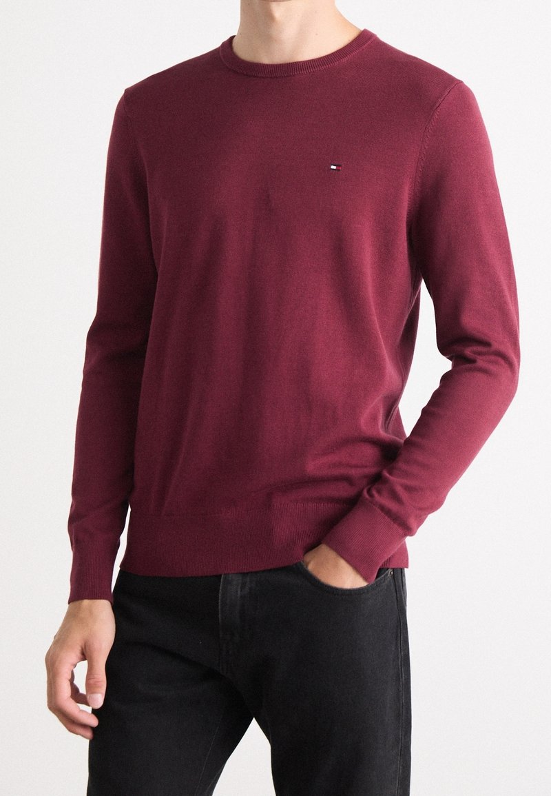 Mann trägt einen taillierten weinroten Rundhalsausschnittpullover mit kleinem Logo auf der Brust, kombiniert mit schwarzen Jeans, eine Hand in der Tasche.