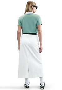 Femme portant un polo vert à manches courtes, une jupe midi blanche avec une fente à l'arrière, des chaussettes blanches, des baskets Nike noires et blanches, et des lunettes de soleil noires, de dos.