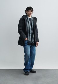 Jachetă neagră impermeabilă, pulover tricotat gri, eșarfă în dungi gri și neagră, blugi albaștri din denim și pantofi slip-on negri, stând în fața unui fundal gri.