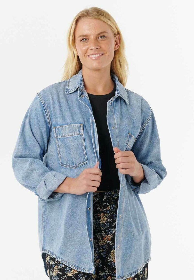 Camisa de mezclilla azul claro con mangas largas, botones a presión en la parte frontal y un bolsillo en el pecho, llevada sobre una blusa negra y pantalones de estampado floral.