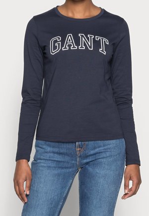 Personne portant une chemise à manches longues bleu marine avec "GANT" en lettres blanches et un jean bleu, visible du cou jusqu'aux hanches sur un fond uni.