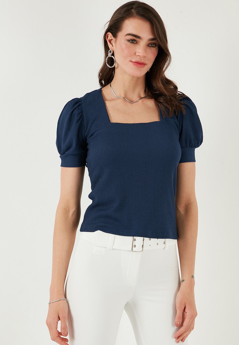 Navy blue textured top with a square neckline and puff sleeves, paired with white pants. Simple design, emphasizing shape and fit.