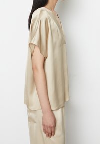 Blouse en soie beige à coupe décontractée, manches courtes et col en V profond. Texture lisse et design minimaliste.