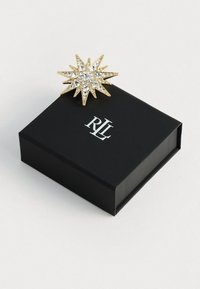 Zlatý brošňa v tvare slnečného lúča, zdobená priehľadnými rhinestones, umiestnená na čiernej škatuľke s bielym logom RL. Škatulka je štvorcová s lesklým povrchom.
