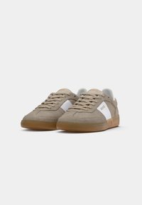 Beige semskinn sneakers med hvite sideakenter og brune gummisåler. Har flate lisser og merkevare på tungen og sidepanelet.
