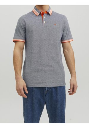 Mand iført grå polo med orange kant og blå jeans står mod ensfarvet baggrund.