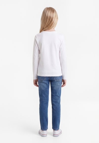 Child with long blonde hair wearing a white long-sleeve shirt, blue jeans, and white sneakers, standing facing away on white background.
