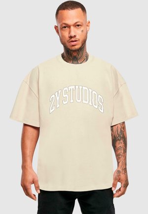 Beige oversized t-shirt with "2Y STUDIOS" printed in large white arch lettering. Features short sleeves and a round neckline.