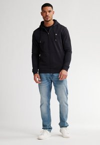 Schwarze Kapuzenjacke mit Reißverschluss und Kordeln, mit einem kleinen Logo. Kombiniert mit hellblauen Jeans und weißen Sneakers.
