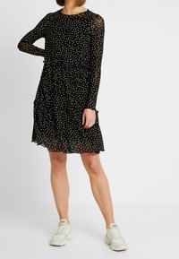 Musta pilkullinen mekko, jossa on pitkät läpikuultavat hihat, kerroksellinen muotoilu ja röyhelöhelma. Yhdistetty vaaleat tossut.