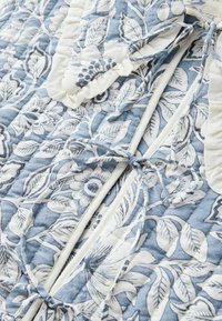 Tessuto trapuntato in un motivo floreale blu e bianco con bordo arricciato e legature decorative. Presenta delicati design di foglie e fiori.