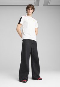 Camiseta blanca de manga corta con acentos negros en los hombros, logo de Ferrari, combinada con pantalones anchos negros y zapatos con detalles rojos.